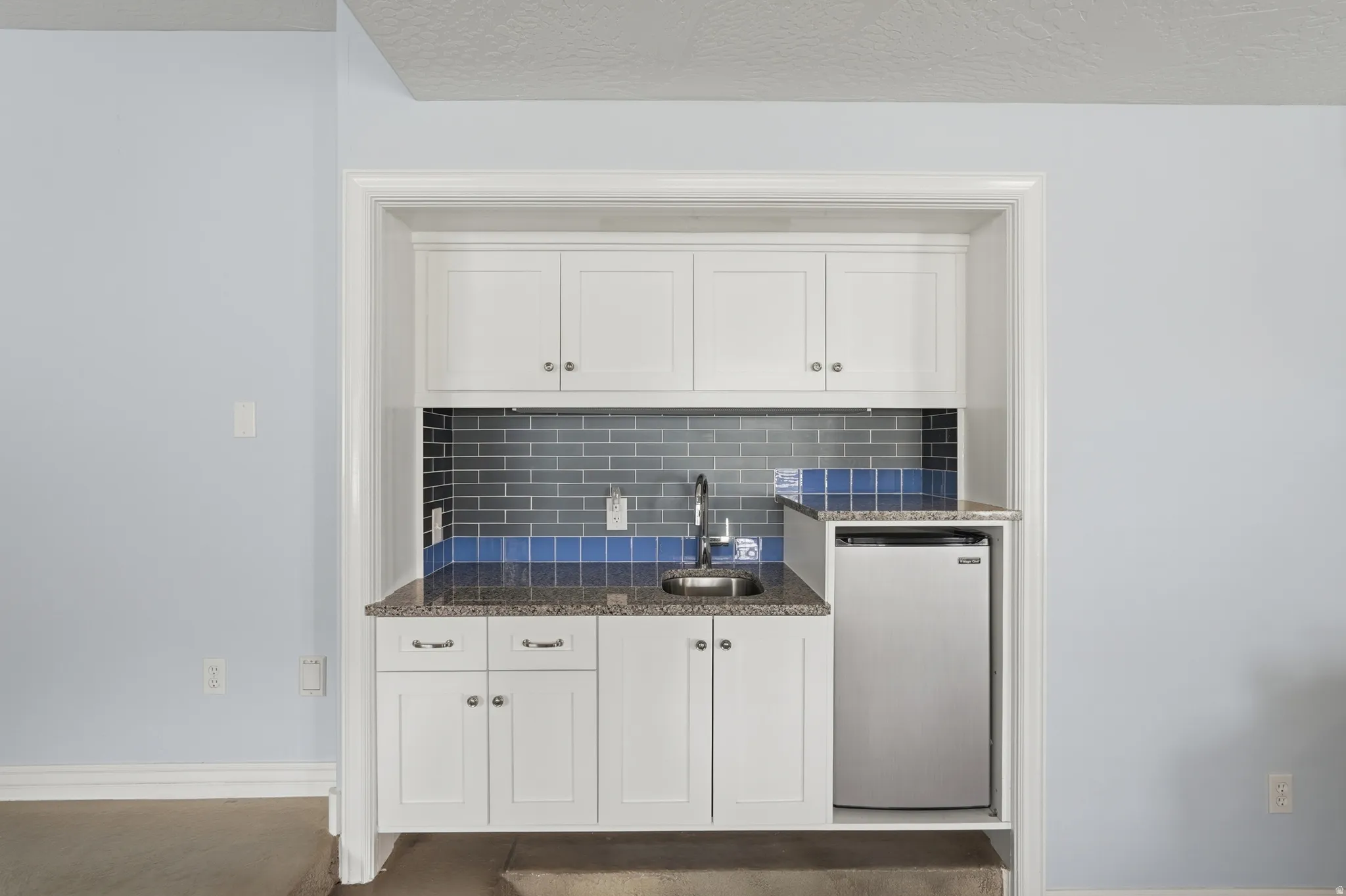 Built-in wet bar featuring white cabinetry, a stainless steel sink, granite-style countertop, and a tiled backsplash