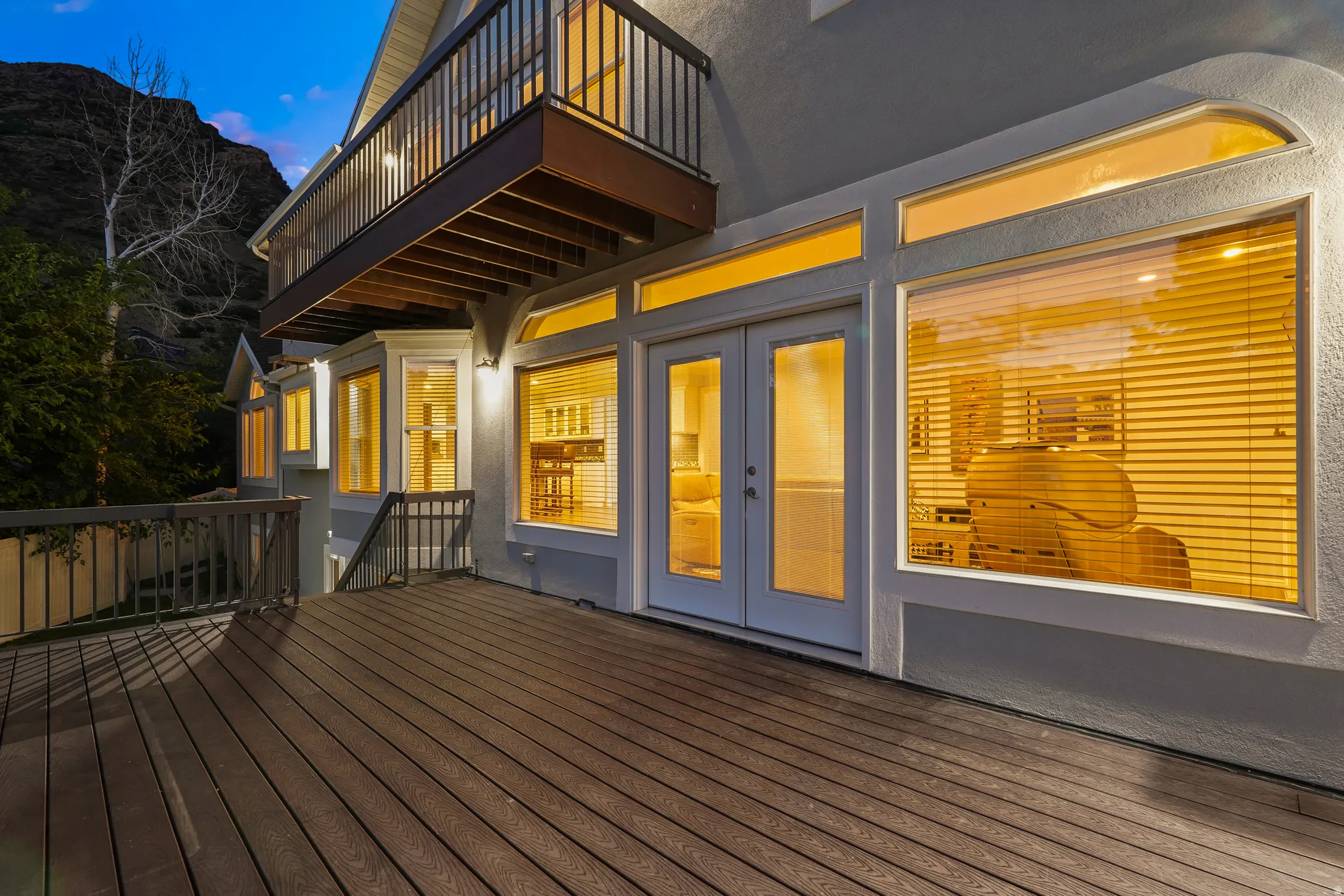 Expansive outdoor deck with wood-finish flooring, featuring a full glass panel double door entry, multiple windows, and an upper-level balcony