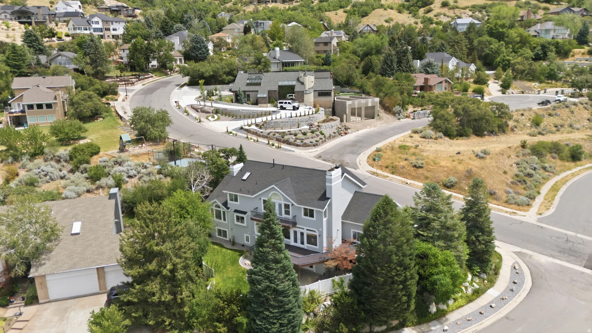 Two-story property featuring a light gray exterior, dark shingle roof, multiple windows, and a surrounding of mature trees and landscaping