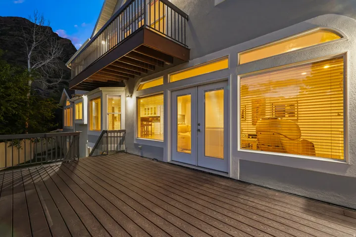 Expansive outdoor deck with wood-finish flooring, featuring a full glass panel double door entry, multiple windows, and an upper-level balcony