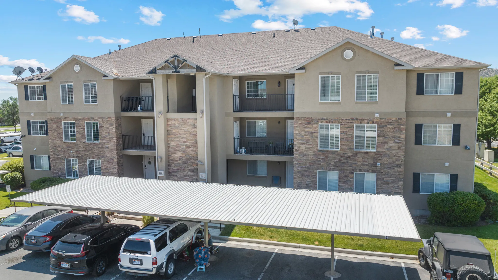 Multi-story building with stone and stucco exterior, covered parking structure, individual balconies, and dark-paneled window shutters