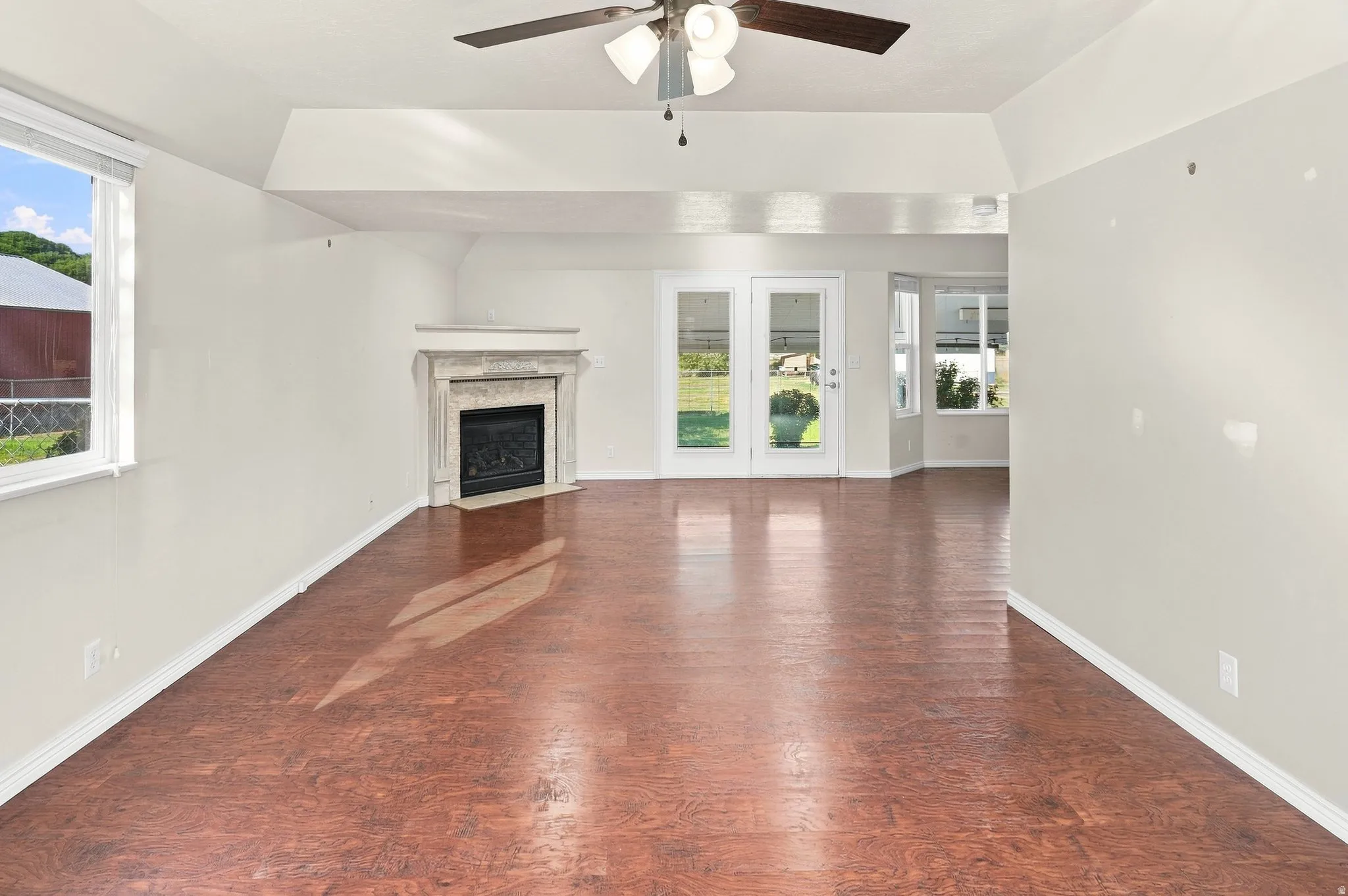 Wood-finish flooring, vaulted ceiling, central fireplace with stone surround, ceiling fan, and French doors