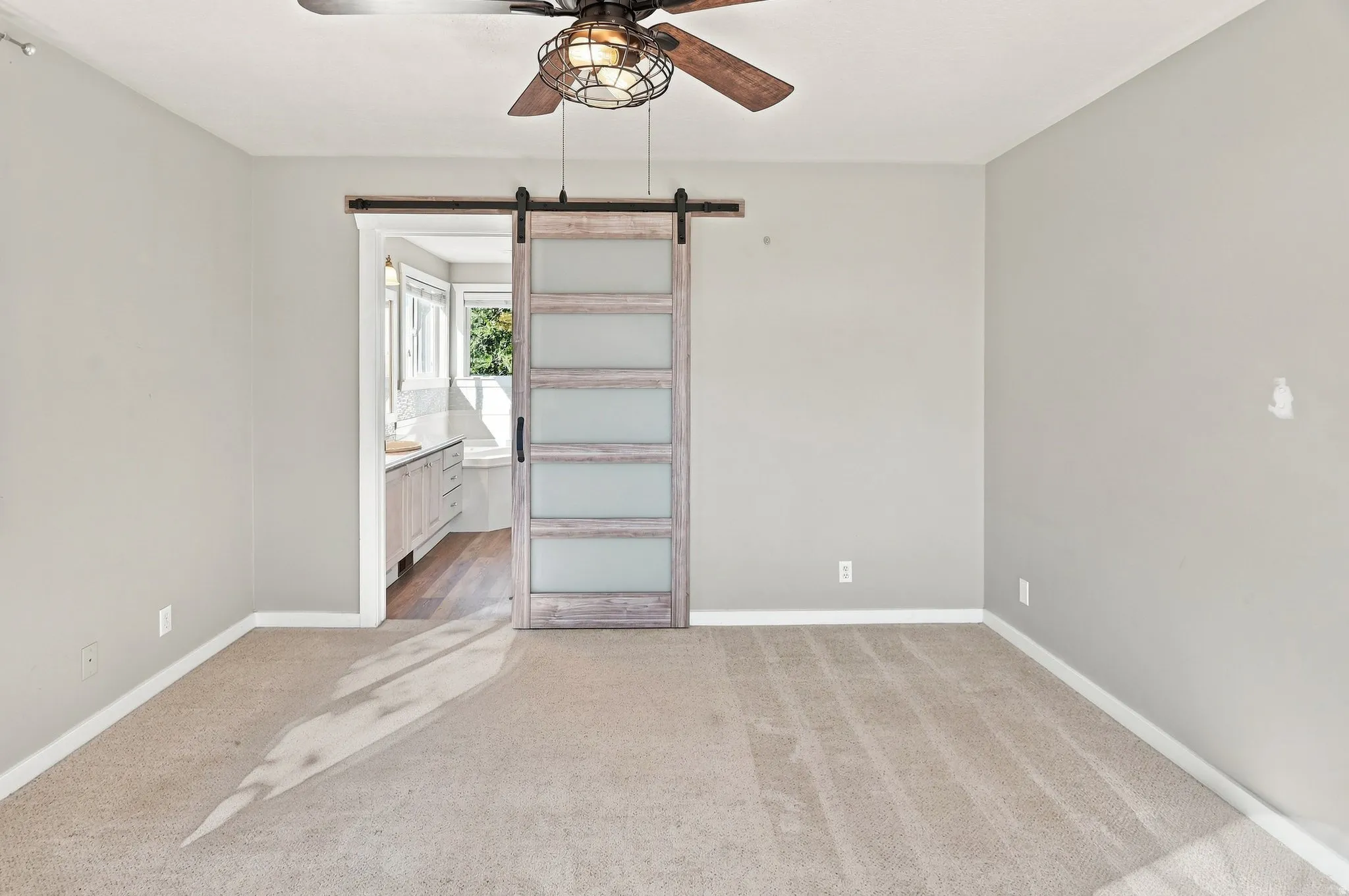 Carpeted flooring, ceiling fan with integrated lighting, a sliding barn door with frosted glass panels, and an adjoining space with wood-finish flooring and windows
