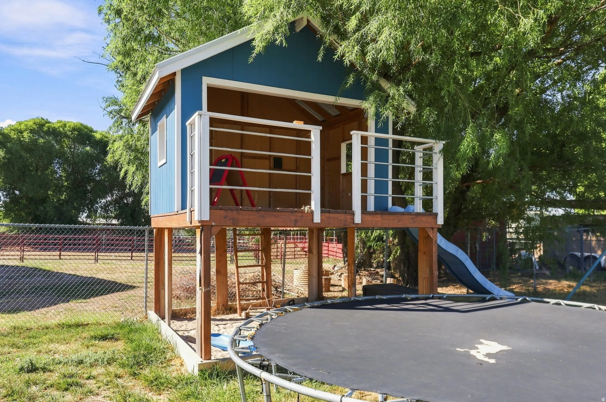 Elevated playhouse with wood framing and blue siding, featuring a covered deck area with white railings and a built-in slide