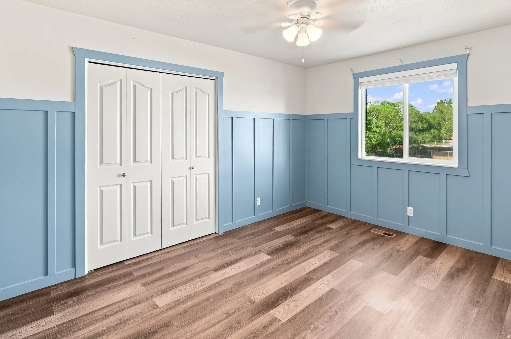 Wood-finish flooring, accent wall with wainscoting, white bi-fold closet doors, and a ceiling fan