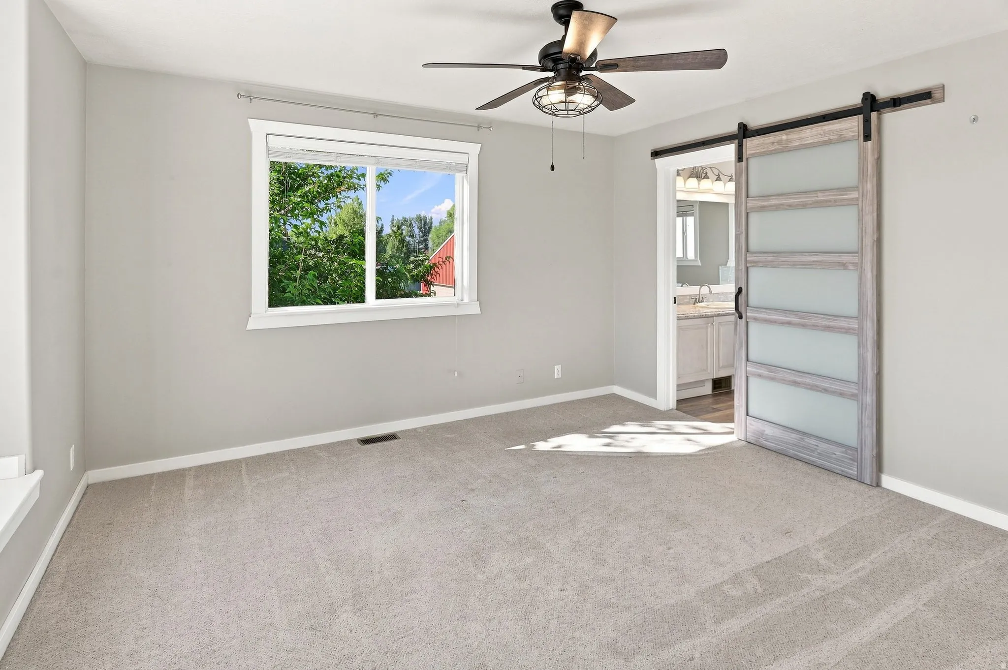 Light gray wall paint, light gray carpeting, ceiling fan with integrated lighting, wood-finish barn door with frosted glass panels, and a window with white trim