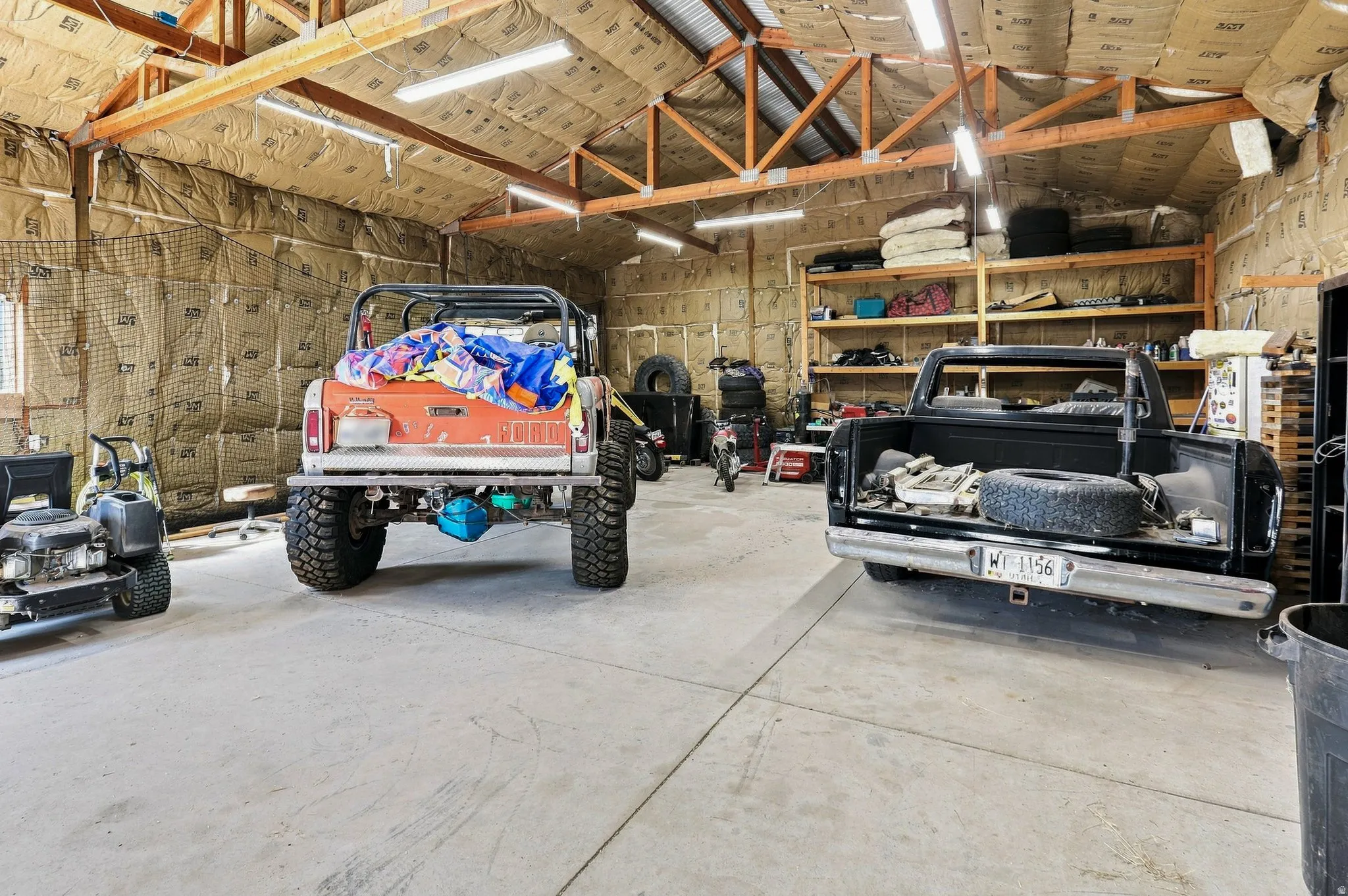 Spacious garage with exposed wood trusses, ample overhead lighting, concrete flooring, and built-in shelving