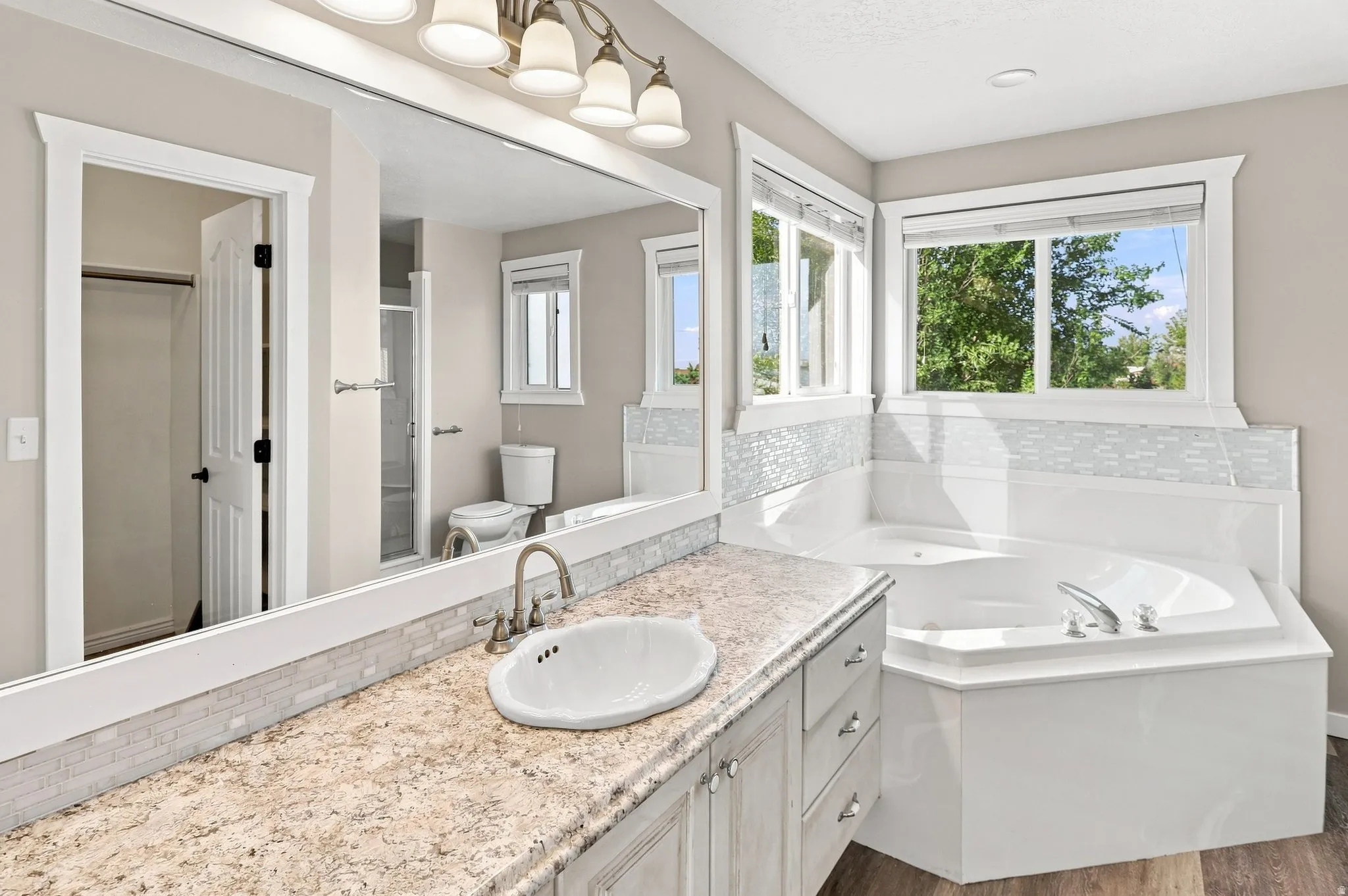 Dual-sink vanity with a light-toned countertop, mosaic tile backsplash, built-in bathtub, and an expansive mirror