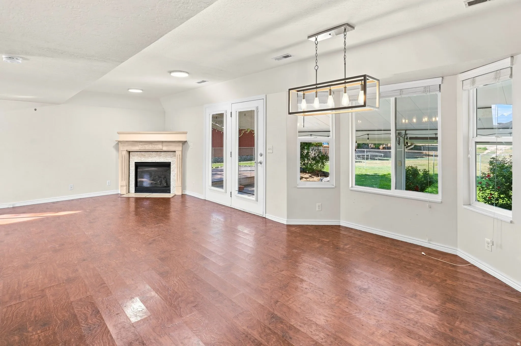 Wood-finish flooring, a fireplace with a stone surround, double French doors, a linear chandelier, and multiple windows