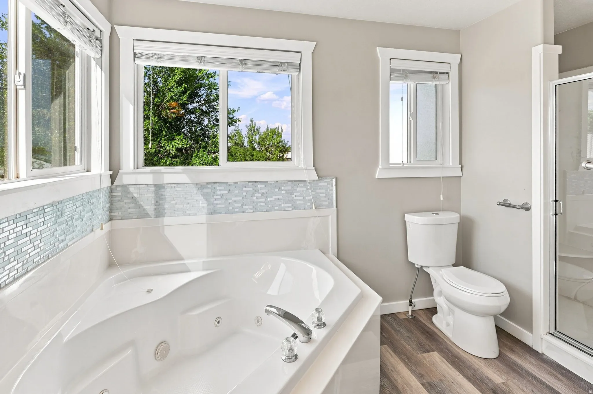 Corner soaking tub with polished chrome fixtures, glass mosaic tile surround, and wood-finish flooring