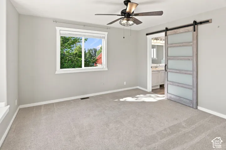 Light gray wall paint, light gray carpeting, ceiling fan with integrated lighting, wood-finish barn door with frosted glass panels, and a window with white trim