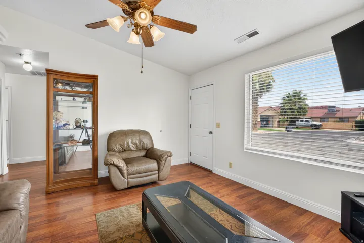 Wood-finish flooring throughout, bright white walls, a large window with horizontal blinds, and a ceiling fan with integrated lighting