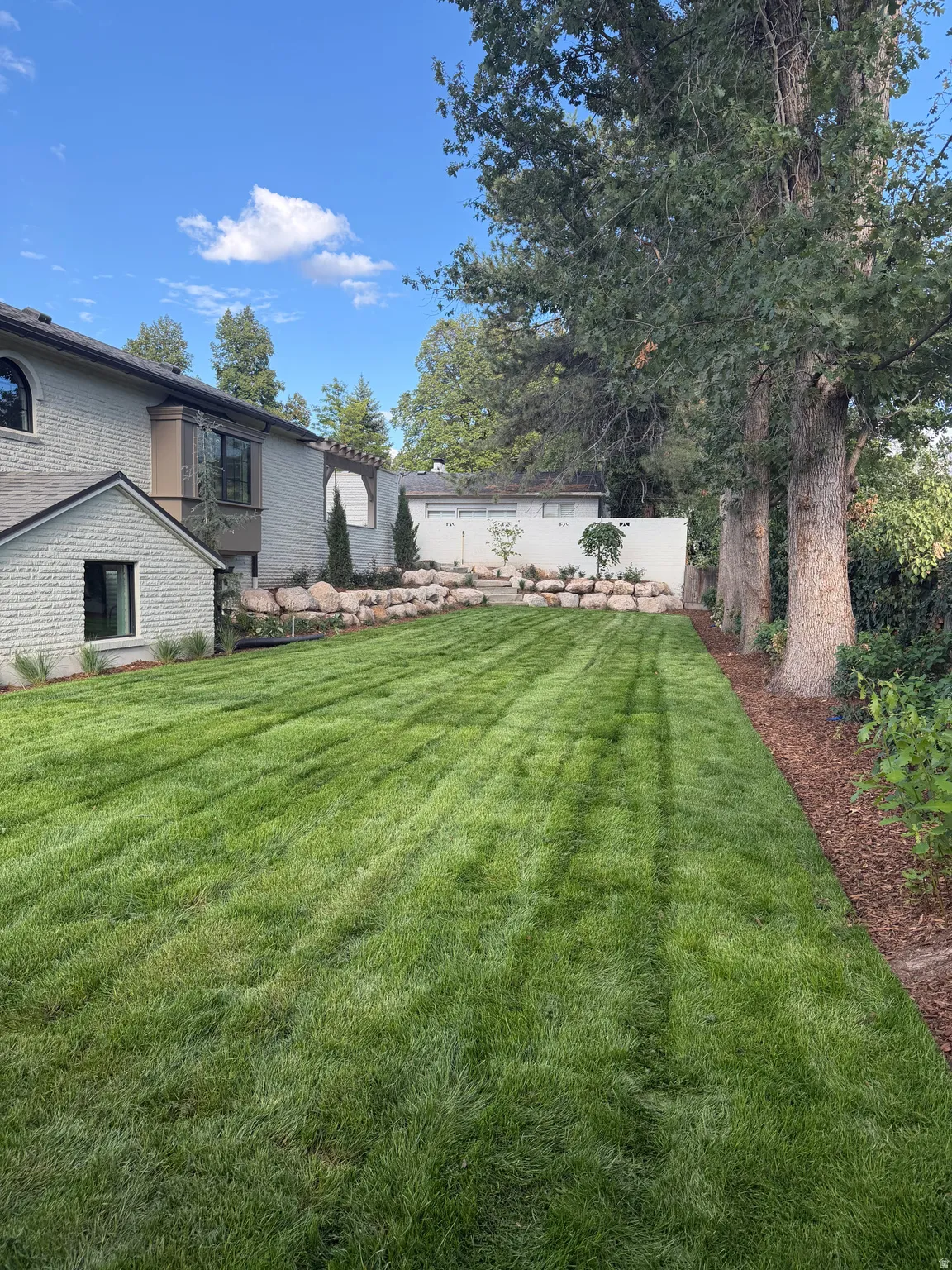 Expansive lawn area with a natural rock retaining wall, mature trees, and a stucco exterior with accent windows