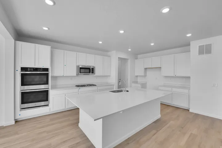 Recessed lighting illuminates the kitchen featuring white cabinetry, light-toned wood-finish flooring, a central island with an undermount sink, and stainless steel built-in double ovens
