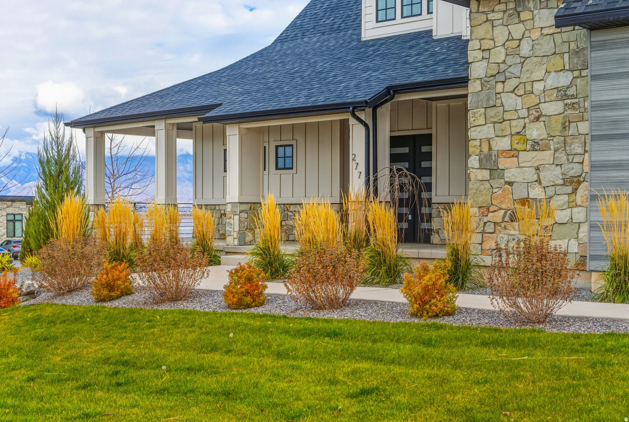 White vertical siding, dark roof, stone veneer accents, covered porch with columns, and a landscaped yard