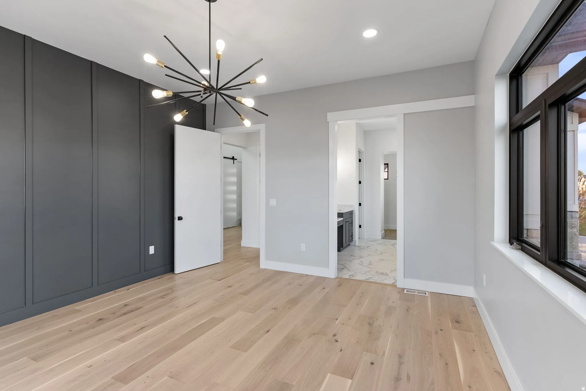 Light wood-finish flooring, accent wall with vertical paneling, modern sputnik chandelier, large window with dark trim, and a pocket door opening to a tiled area