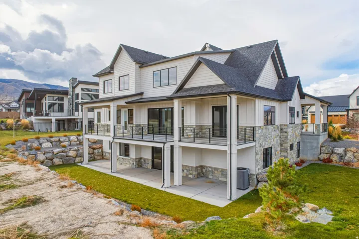 Multi-story residence featuring white siding, stone accents, black-framed windows, multiple balconies with black railings, and a lower-level patio