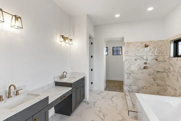 Dual sink vanity with dark cabinetry and brass fixtures, white countertops, large format tile flooring, and a tiled shower with brass fixtures