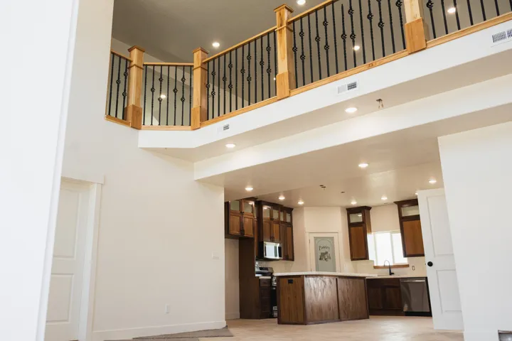 Open-concept interior featuring a second-story balcony with wood and wrought iron railing, recessed lighting throughout, and a kitchen with wood cabinetry, a central island, and stainless steel appliances