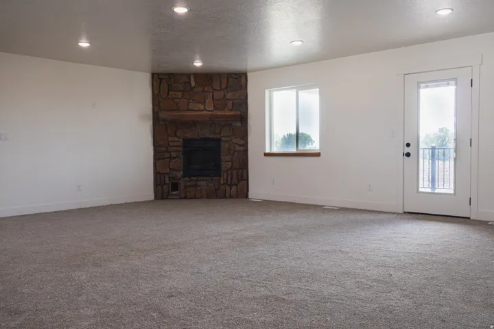 Neutral-toned carpeting throughout, stone-clad fireplace with wood mantel, recessed ceiling lighting, and abundant natural light from a window and glass-paned exterior door