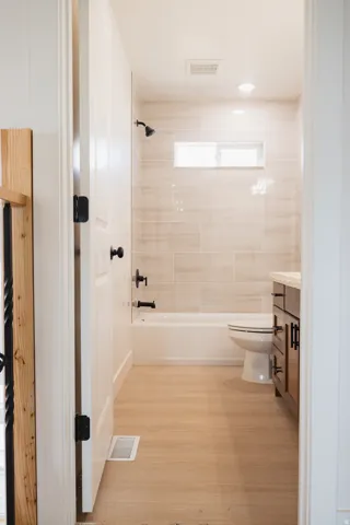 Wood-finish flooring, recessed lighting, tile shower surround, window above the bathtub, and dark wood vanity with a light-colored countertop
