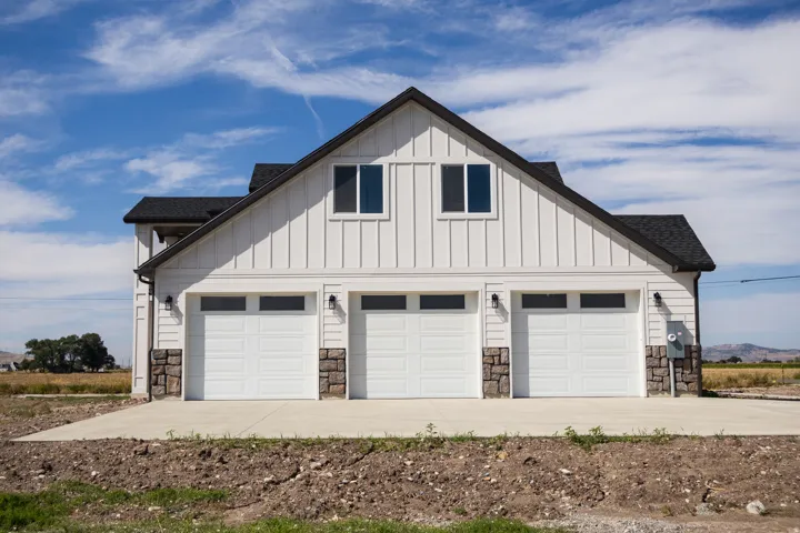 Three-car garage with white panel doors and upper window panels, board and batten siding, stone accents, black asphalt shingle roof, and exterior lighting fixtures