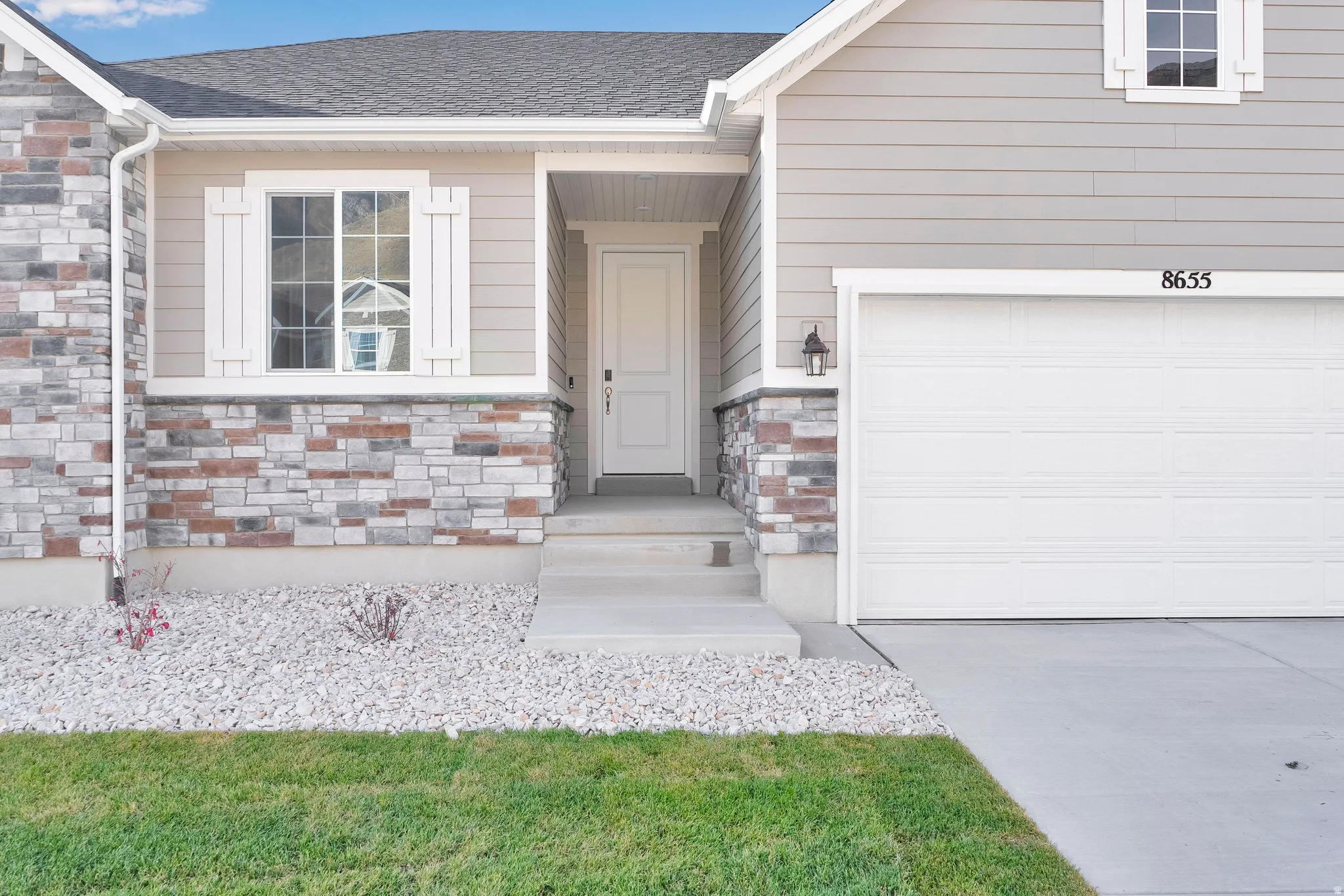 Stone veneer detailing, light gray horizontal siding, white window trim with shutters, single-panel garage door, and concrete front entry
