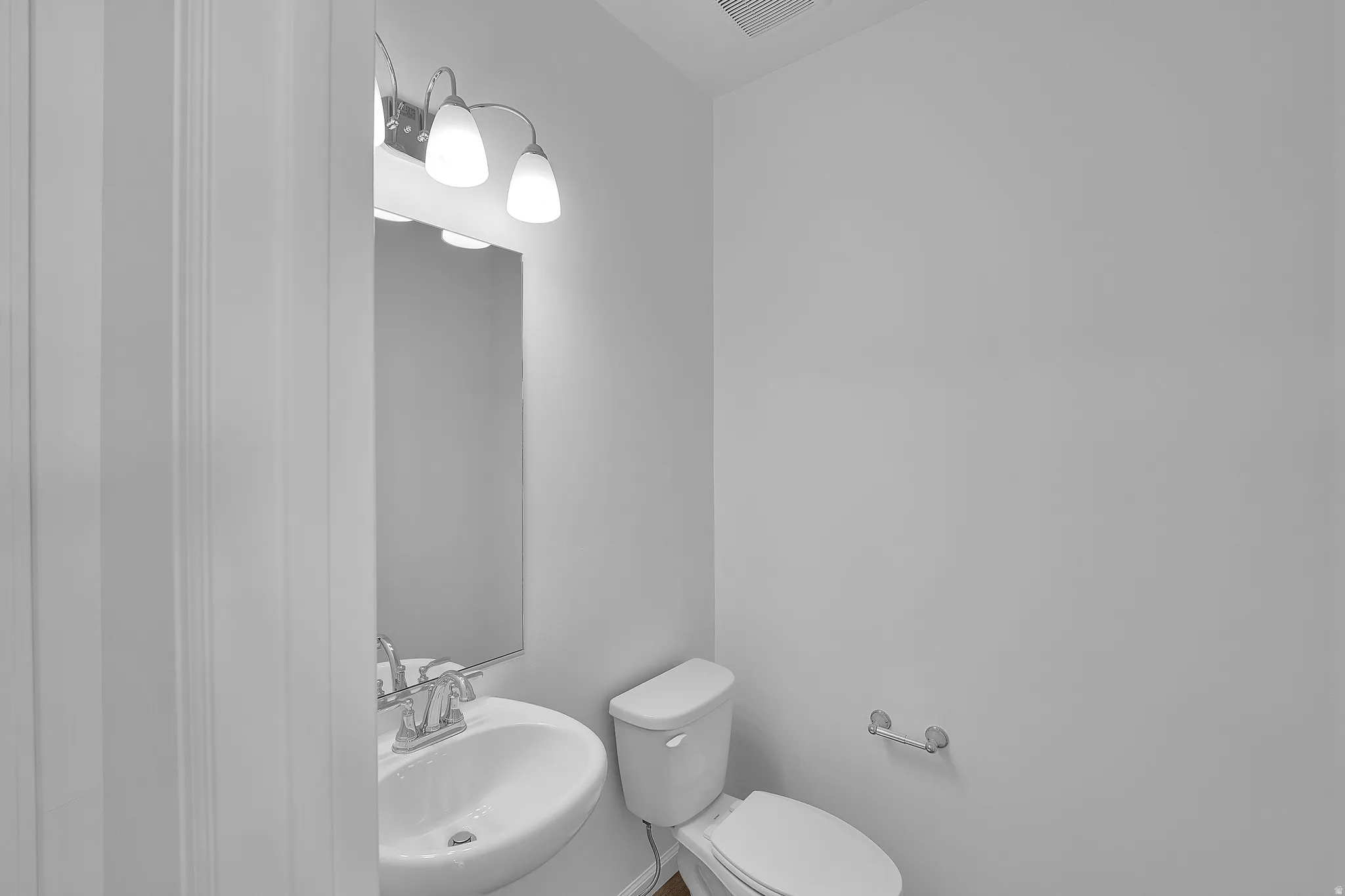 Wall-mounted vanity lighting, mirror, pedestal sink with chrome faucet, toilet, and wall-mounted toilet paper holder
