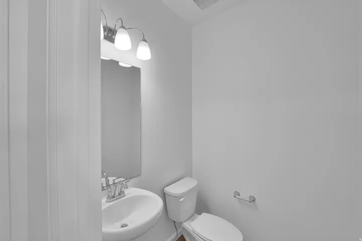Wall-mounted vanity lighting, mirror, pedestal sink with chrome faucet, toilet, and wall-mounted toilet paper holder