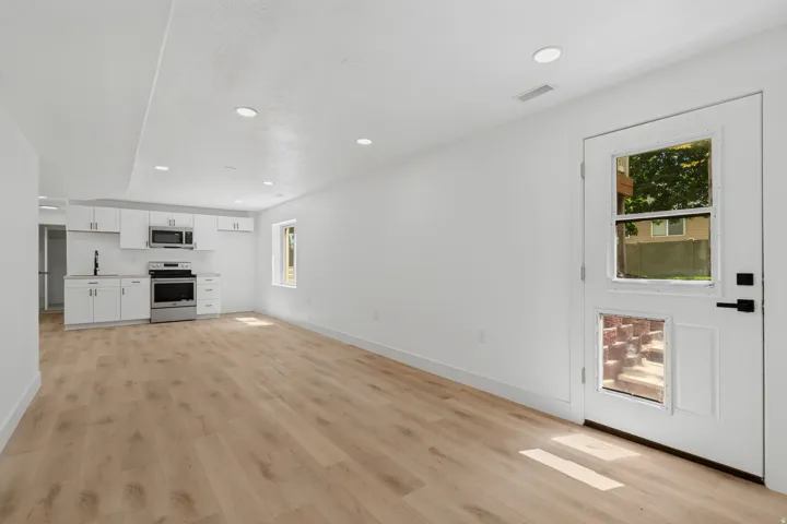 Recessed lighting illuminates the open-concept space featuring wood-finish flooring and a kitchen with white cabinetry, stainless steel appliances, and a window