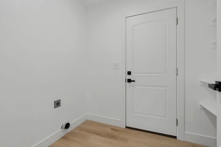 Laundry area with wood-finish flooring, white walls, and a paneled door with matte black hardware