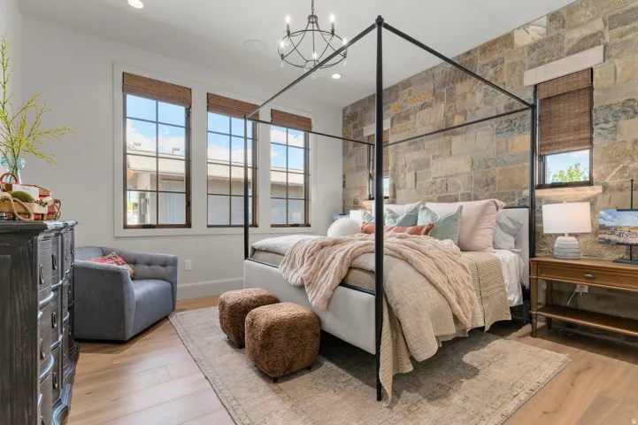 Stone accent wall, wood-finish flooring, three grid-pattern windows with woven blinds, and a wrought iron chandelier