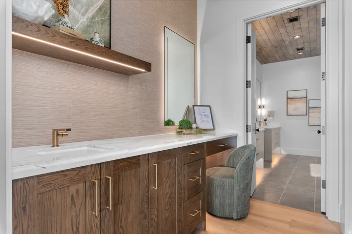 Vanity area featuring a built-in wood-finish cabinet with gold-tone hardware, white countertop, integrated sink, and an illuminated floating shelf