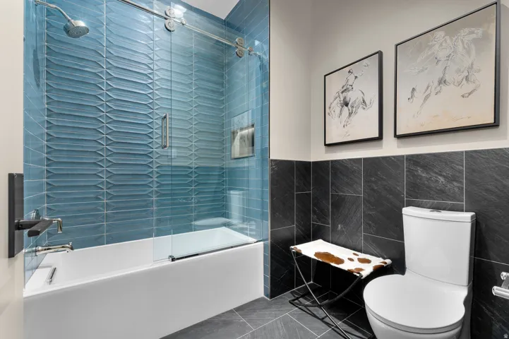 Built-in bathtub with a sliding glass enclosure, blue textured tile surround, wall-mounted showerhead, and dark large-format tile flooring