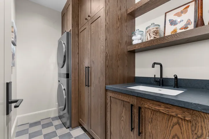 Stacked washer and dryer, wood-finish cabinetry, built-in shelving, undermount sink with a matte black faucet, and checkerboard floor tiling