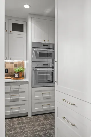Light gray cabinetry with gold-tone hardware, stainless steel built-in appliances, patterned floor tiling, and a textured backsplash
