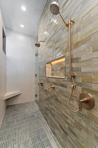 Expansive shower featuring dual copper-toned shower heads, linear tile flooring, a built-in bench, a recessed niche with integrated lighting, and a glass partition