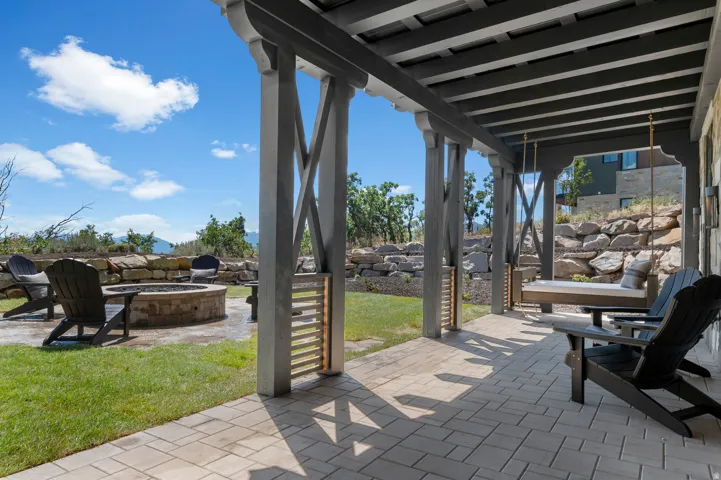 Expansive outdoor living area featuring a paved patio, a built-in fire pit, a covered pergola, and a stone retaining wall