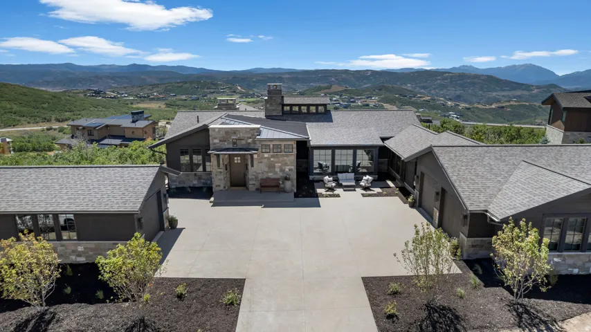 Modern exterior with stone and wood siding, expansive concrete driveway, and multiple rooflines, set against a mountainous backdrop