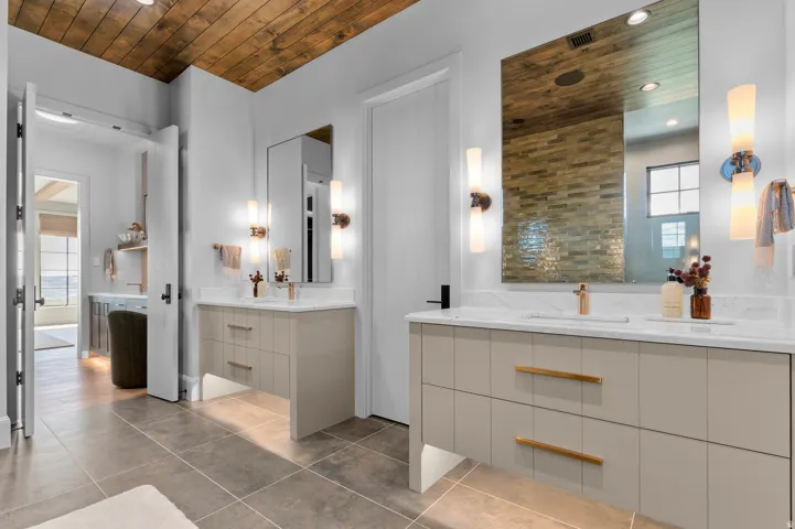 Wood-paneled ceiling, dual floating vanities with light cabinetry, stone countertops, and integrated rectangular sinks, complemented by large wall mirrors