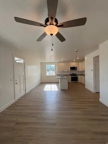 Open-concept living space featuring wood-finish flooring, a ceiling fan with integrated lighting, white walls, and recessed lighting in the kitchen area