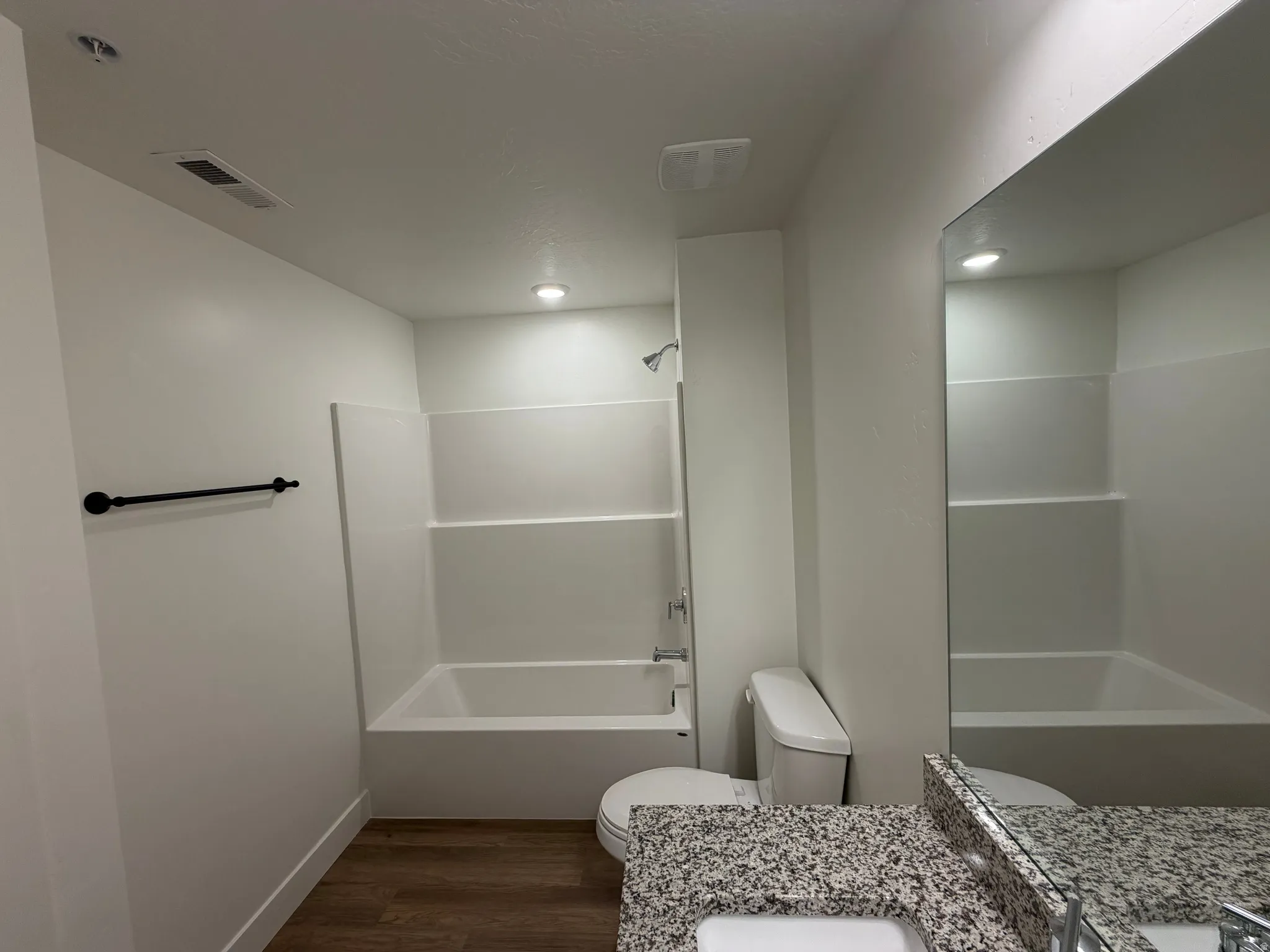 White bathtub with integrated shower, wood-finish flooring, and a granite-patterned vanity countertop with an undermount sink