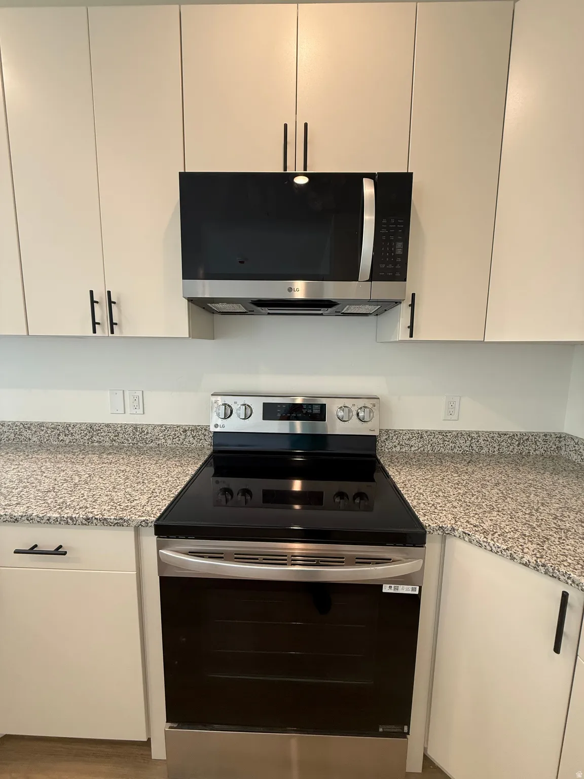 Stainless steel range with a black ceramic cooktop, coordinating over-the-range microwave, white upper and lower cabinetry with matte black hardware, and speckled light-toned countertops
