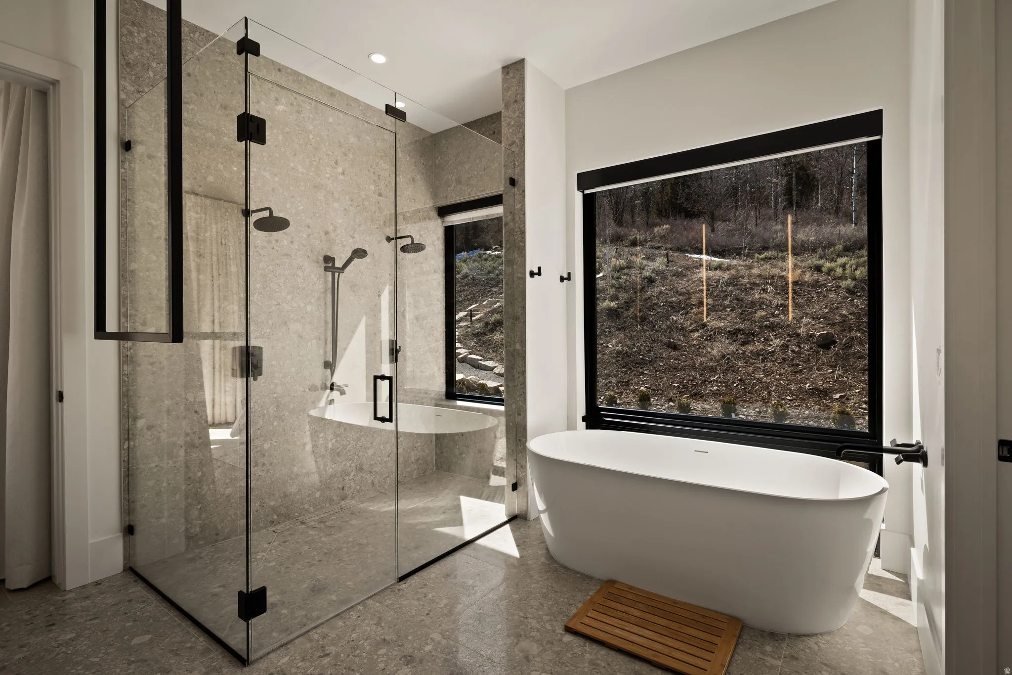 Freestanding soaking tub with a large window, a glass-enclosed shower with dual showerheads, and neutral-toned tile flooring and walls