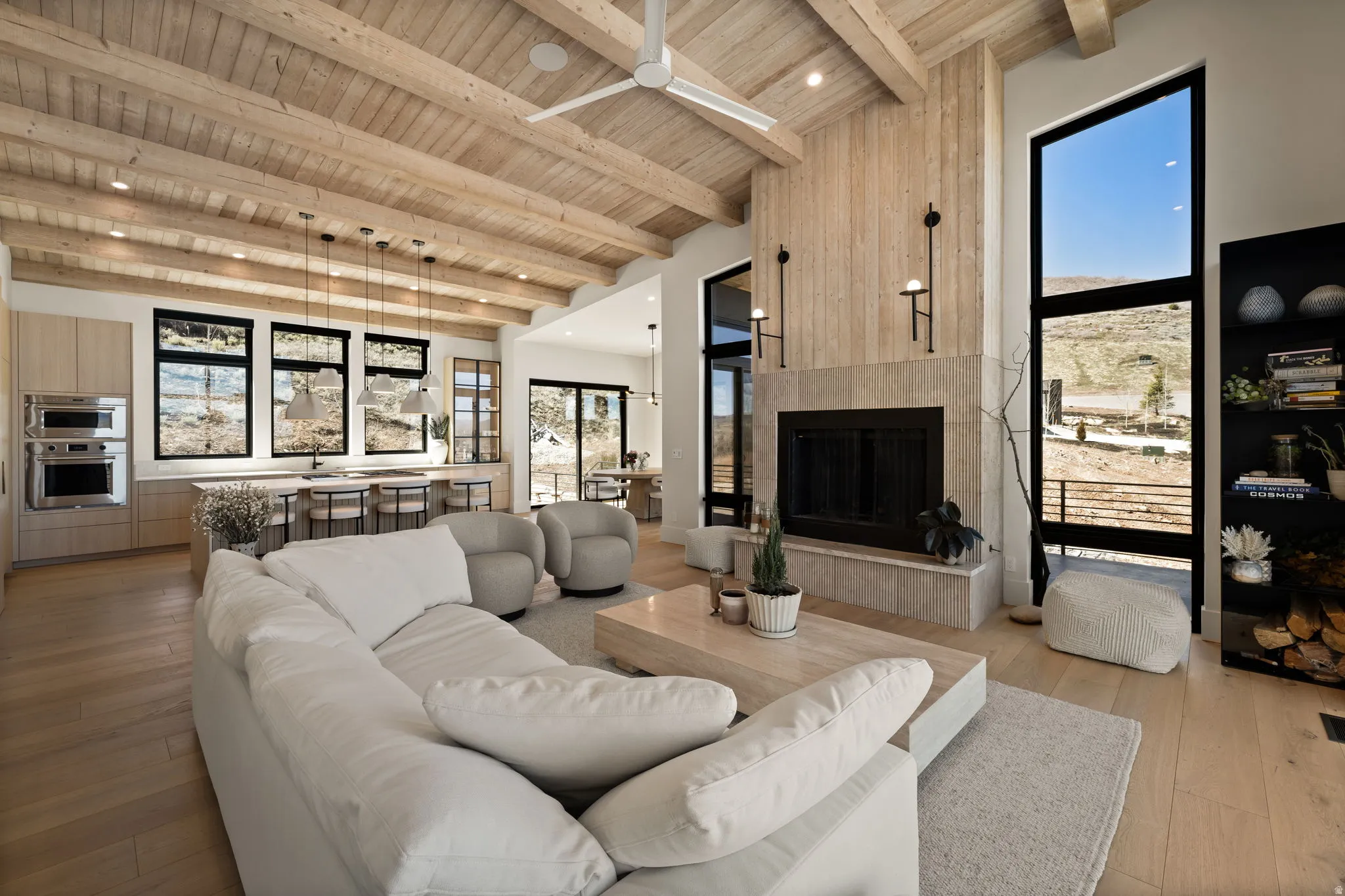 Vaulted wood-clad ceilings with exposed beams, stone-clad fireplace with wood paneling, floor-to-ceiling windows, light wood-finish flooring, and built-in kitchen appliances