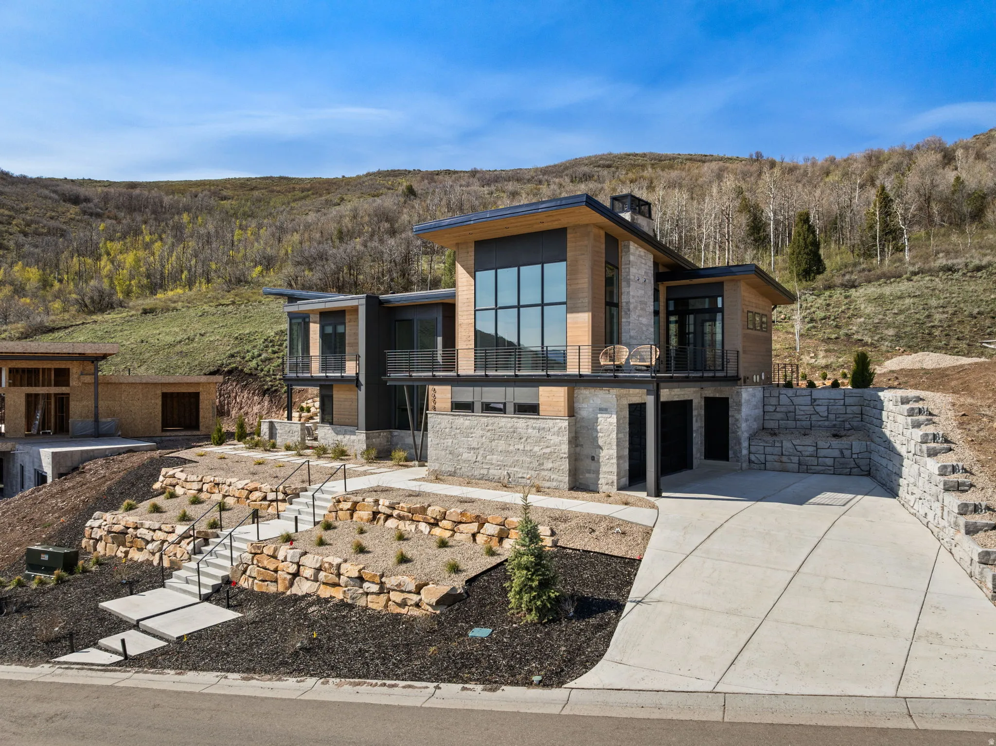 Modern architectural design with a multi-level structure, stone and wood-panel exterior, expansive window walls, an integrated garage, and a concrete driveway