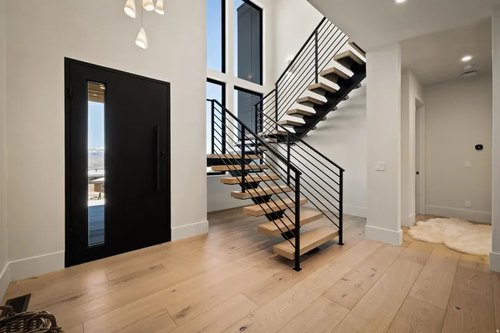 Modern entryway featuring a multi-paneled front door with vertical glass insert, light wood-finish flooring, a floating staircase with black metal railings, and a two-story window