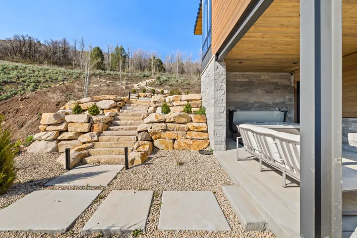 Stone retaining walls with integrated landscaping, stone staircase, and concrete pad pathway leading to a covered patio