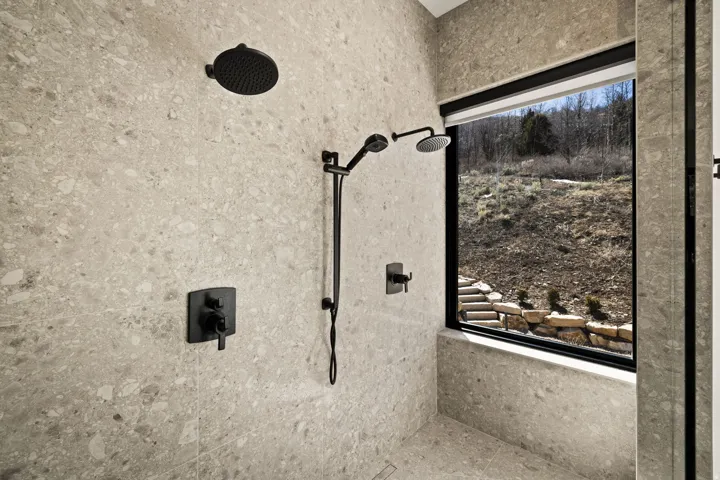 Expansive shower with speckled tile walls, dual matte black shower heads, and a large window offering natural light