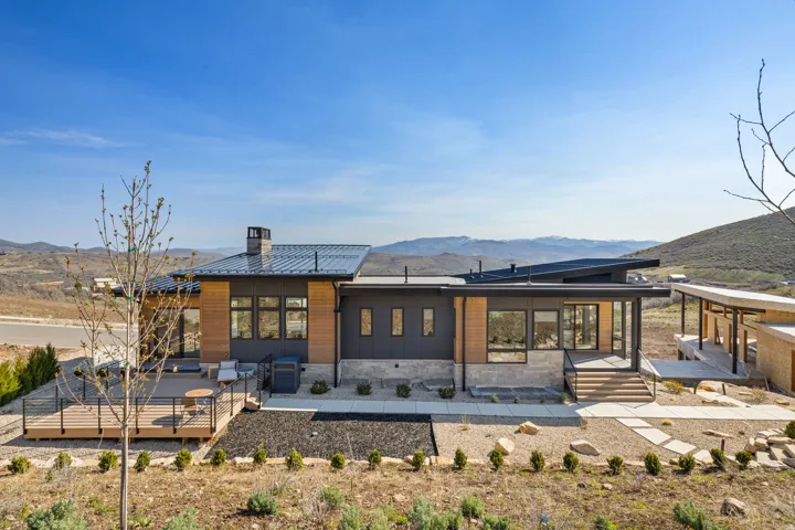 Modern exterior featuring a dark metal roof, wood siding accents, expansive windows, a spacious wood deck, and a stone retaining wall